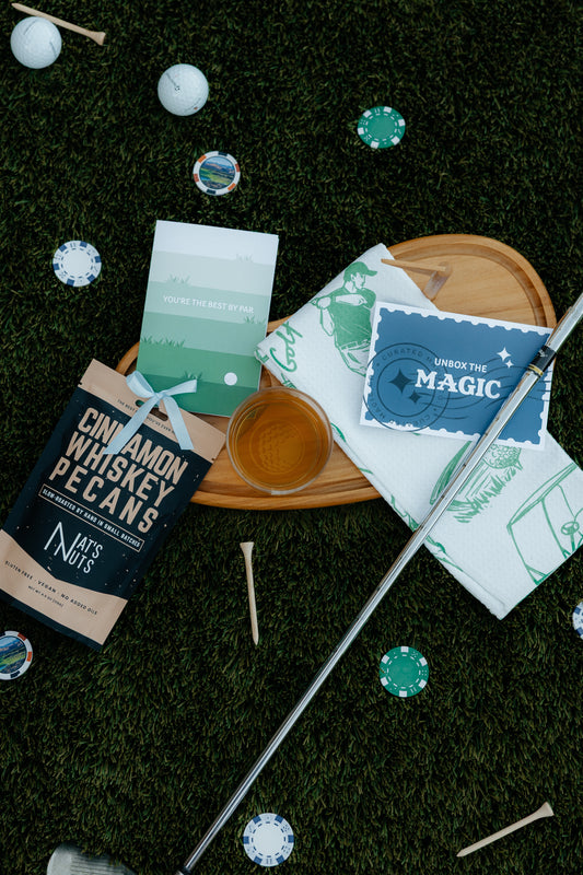 Golf Themed charcuterie board with cinnamon whiskey pecans, golf towel, best by par card and drink. Card saying unbox the magic