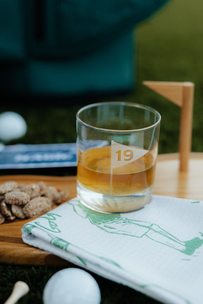 Glass of amber liquid on a golf-themed towel with a blurred golf course background