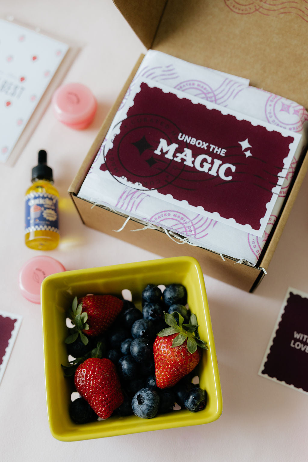 Box with 'Unbox the Magic' text, bottle, and container of strawberries and blueberries on a light surface.
