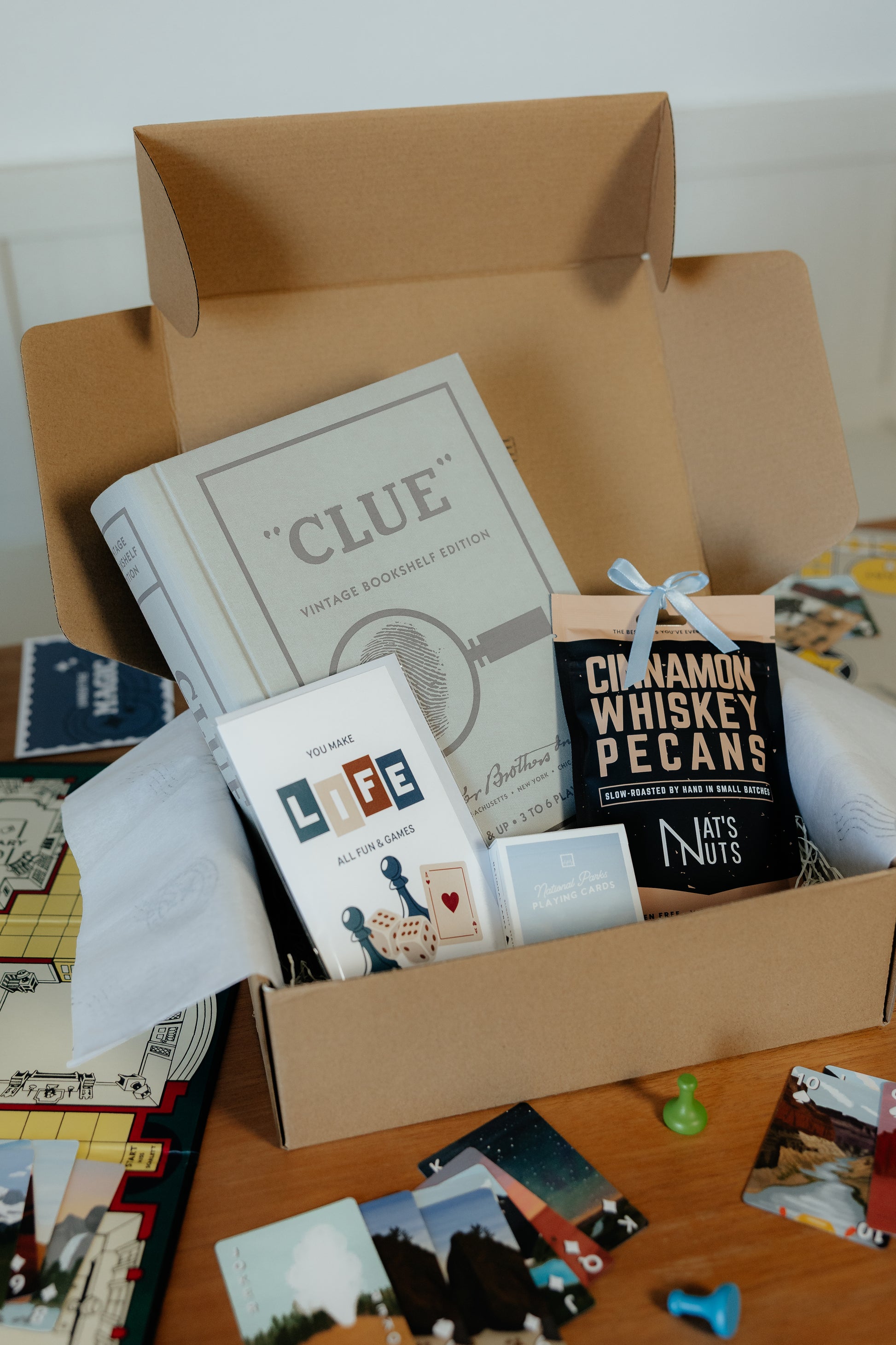 Open cardboard box containing 'Clue' game book, greeting card, cinnamon whiskey pecans and playing cards. 