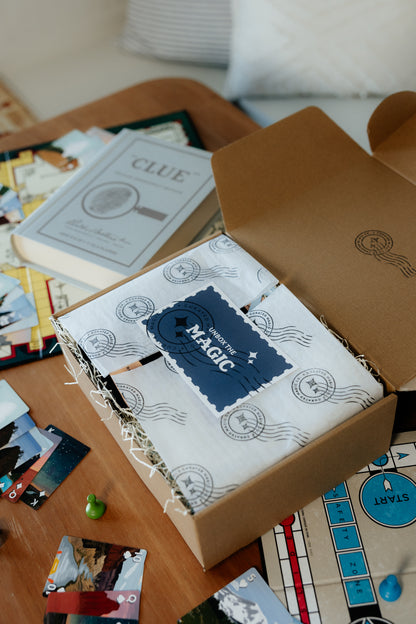 Open box with a  card 'Unbox The Magic' on a table with board game pieces and books.