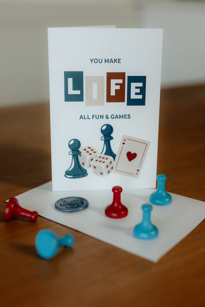 Greeting card with text "you make life all fun & games" with game pieces on a wooden surface